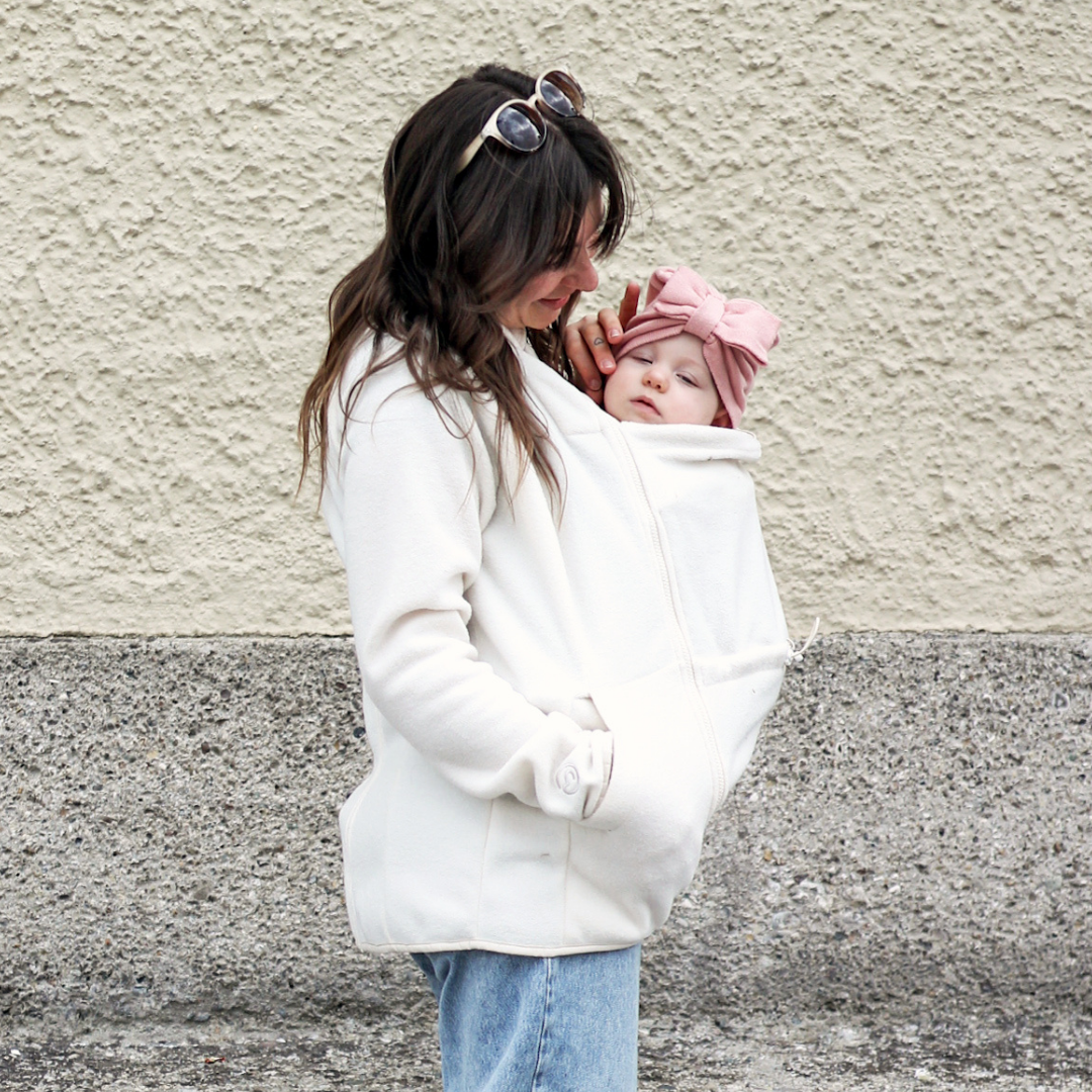 Mutter trägt Baby unter heller Fleece-Tragejacke, beide eng eingekuschelt, ruhige Szene