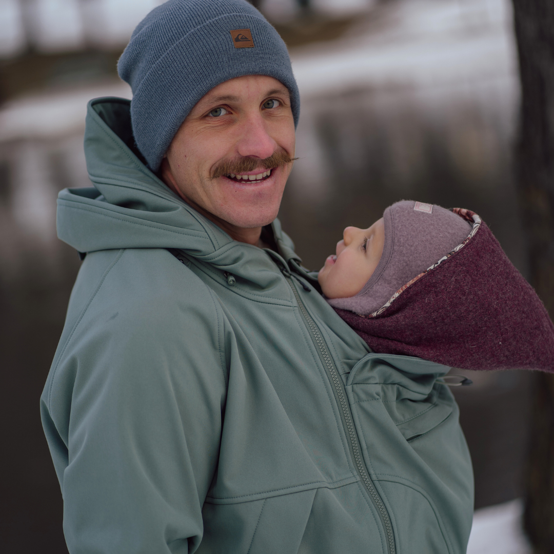 Vater trägt Baby in grüner Softshell-Tragejacke, beide warm gekleidet, winterliche Umgebung