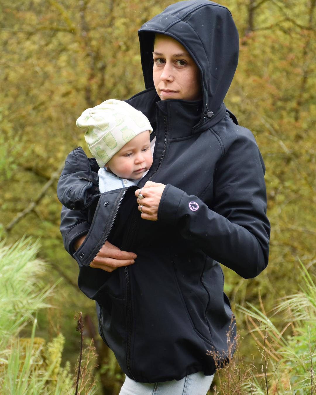 Frau trägt Baby unter schwarzer Jacke mit Kapuze in herbstlicher Natur
