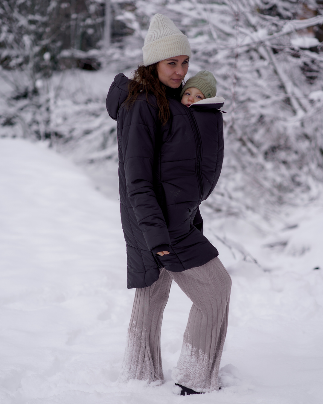 Mutter trägt Baby in dunkler Stepp-Tragejacke im Schnee, winterliche Landschaft, beide warm gekleidet