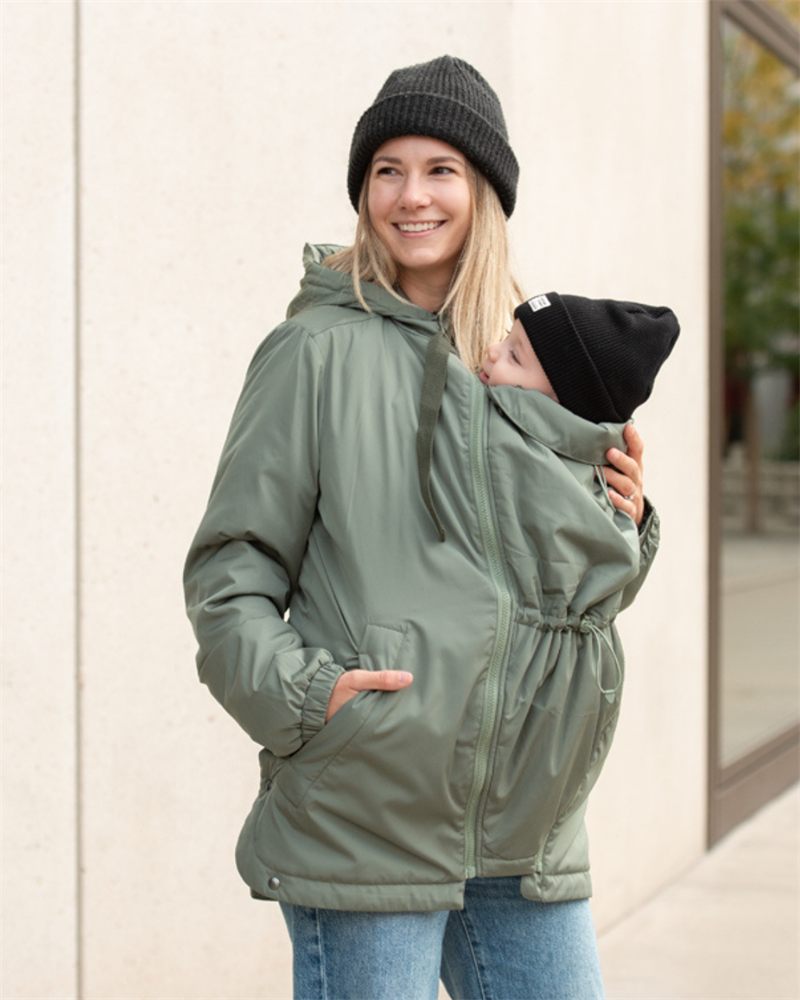 Mom carrying her baby in a green mamalila babywearing jacket in front of a wall