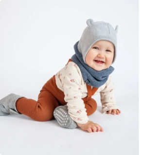 Ein glückliches Baby mit grauer Mütze und blauen Schal krabbelt am Boden vor neutralem Hintergrund.
