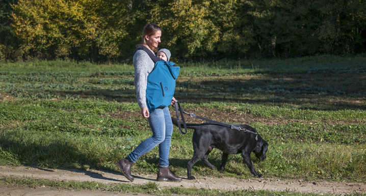 Mutter geht mit Kind im Tragecover und Hund spazieren